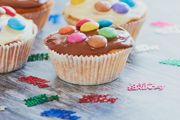 selfmade muffins coated with chocolate and chocolate lentils on wooden surface beside small birthday decoration with the words: happy birthday