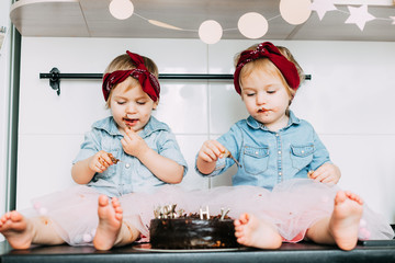 two little twin girls celebrate their birthday. eating chocolate cake while sitting in the kitchen. stylishly dressed in denim shirts and tulle skirts