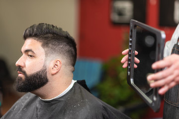 Handsome bearded man looking in the mirror after haircut, at barber shop .