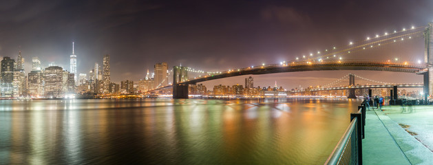 Fototapeta na wymiar A panorama of the Brooklyn Bridge at night with the New York City skyline in the background. The East River is in front and parts of the Manhattan Bridge can be seen.