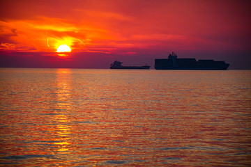 Small and big container tanker ship silhouette parked in a bay with a beautiful sunset sky and reflection in the ocean sea