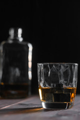 Vertical photograph of a glass of whiskey with unusual edges and ice stones on a wooden table and a bottle in the defocus in the background.