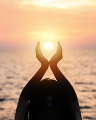 June summer sun solstice concept with silhouette of happy young woman's hands relaxing, meditating and holding sunset against warm golden hour sky on the beach with natural ocean or sea background
