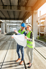 Two young architects or civil engineers visiting large construction site, looking at floor plans. Man is pointing into distance with his hand