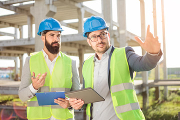 Two young male architects or business partners talking on a construction site. One man is pointing...