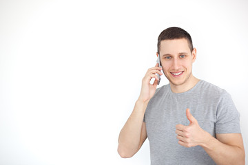 Portrait of a cheerful, positive, attractive guy with bristles in a gray T-shirt, with a black screen smartphone in his hand, showing a thumbs up gesture on white background.  Portrait man 