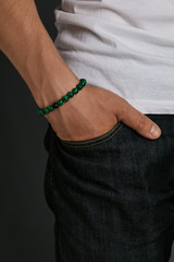 Cropped half-turn shot of guy's hand with stony bracelet with smooth surface on the malachite beads. The man in jeans and T-shirt is putting his hand into pocket, posing against the dark background.