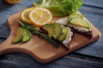Dark burger with grain bread on dark ceramic plate, salad, rosemary and lemon