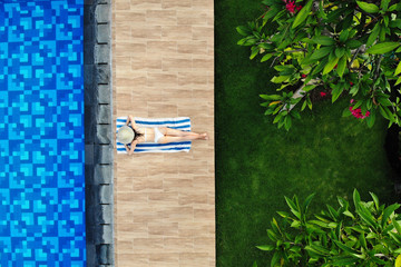 Top view of slim young woman in white bikini and straw hat lying on towel near swimming pool. Back view, without face