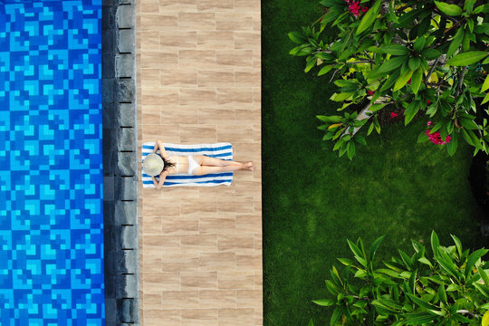 Top View Of Slim Young Woman In White Bikini And Straw Hat Lying On Towel Near Swimming Pool. Back View, Without Face