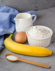 The ingredients for the banana cake on gray background. Wooden Spoon, Banana, Eggs, Milk