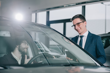 cheerful dealer in glasses standing near curly man in red car