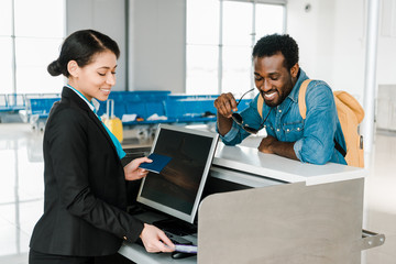 smiling african american airport worker checking air ticket and passport at ticket control in...