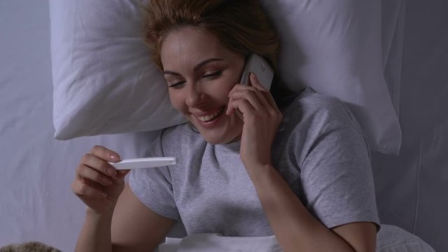 Smiling woman in bed holding pregnancy test and talking phone, positive result