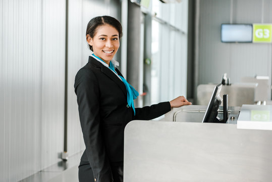 smiling african american airport staff in uniform standing at check in desk and looking at camera