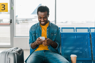 smiling african american man sitting in departure lounge and using smartphone