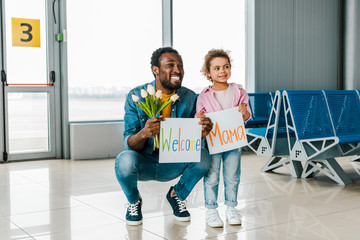 african american daughter and father standing in waiting hall in airport and holding tulips and...