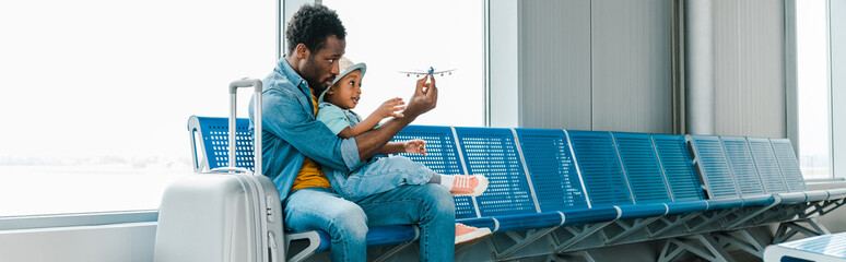 panoramic shot of african american father and son sitting with suitcase in airport and playing with...