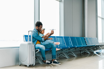 african american father and son sitting with suitcase in airport and playing with toy plane