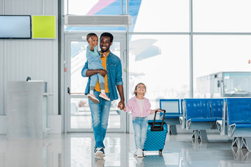 smiling african american father walking with happy children and baggage along waiting hall in...