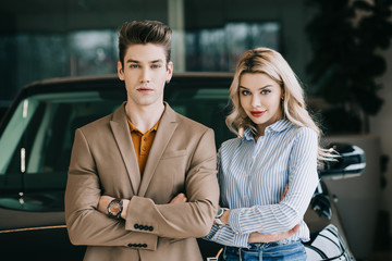 handsome man and beautiful blonde woman standing with crossed arms in car showroom