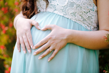 A pregnant woman in a blue dress, depicting a heart with her hands on her stomach.