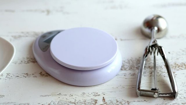Young housewife weighs prunes on a table scales on a white plate on a bright wooden table in the kitchen. Women's hands. Close-up.