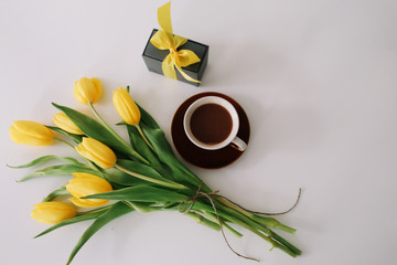Cup of coffee, a gift box with ribbon and bouquet of yellow tulips isolated on white background. Flat lay, top view, copy space