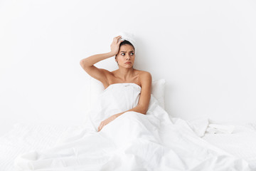 Sad amazing woman with towel on head lies in bed under blanket isolated over white wall background with headache.