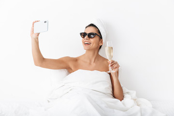 Beautiful emotional woman with towel on head lies in bed under blanket isolated over white wall background wearing sunglasses take a selfie by mobile phone.