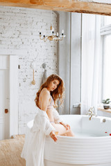 Beautiful girl with long curly hair in sexy underwear posing on bathroom background. Woman sitting on the edge of the bathroom with a glass of champagne