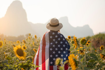 Woman is holding USA flag and walking into Sunflower filed plantation with landscape view as for...