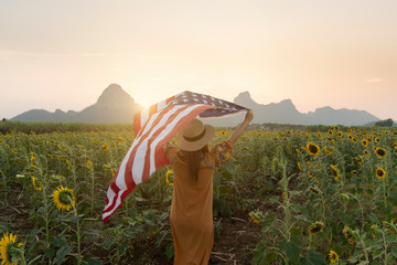 Woman is holding USA flag and walking into Sunflower filed plantation with landscape view as for concept of 4th July - USA national day.