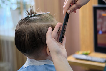 Fototapeta premium Barber shop. Hairdresser makes hairstyle to a boy with hair scissors and black comb. 