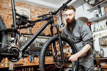 Handsome bearded repairman in workwear mounting wheel on a mountain bicycle at the workshop