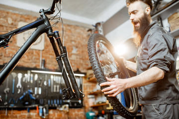 Handsome repairman in workwear serving mountain bicycle, standing with front wheel at the workshop of a bicycle shop