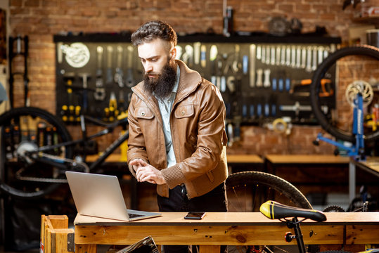 Stylish bearded businessman as a shop owner or manager working with laptop at the bicycle workshop