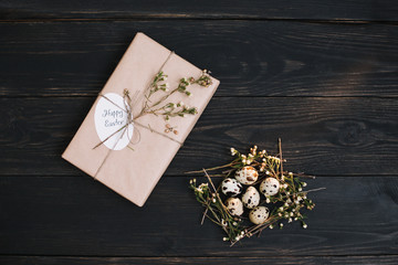 Easter eggs in birds nest on dark wooden background with a gift. Rustic easter still life with quail eggs, dry willow branches on dark background. Flat lay, top view