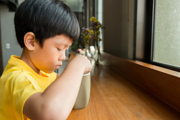a little boy sitting at the wooden table by the window at home to drink a cup of hot chocolate. he hold the cup in his left hand and mixed hot chocolate with cream with a teaspoon in his right hand.