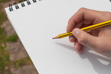 Boy's hand with a pencil over an open notepad in the park.