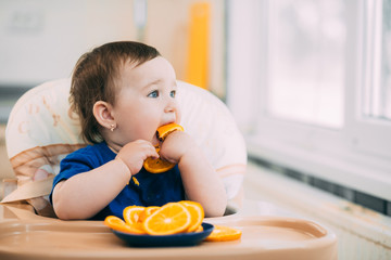 a little girl in a blue t-shirt and a blue plate sitting in a child's chair eating an orange