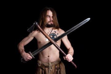 young strong long-haired bearded man with a tattoo and a sword and an ax of the Viking era looks to...