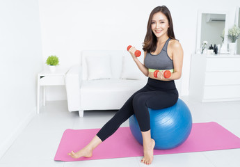 Portrait beautiful Asian young sporty woman working out with dumbbells sitting on gymnastic blue fit ball and looking at camera in home. Sport, fitness,People and healthy lifestyle concept.