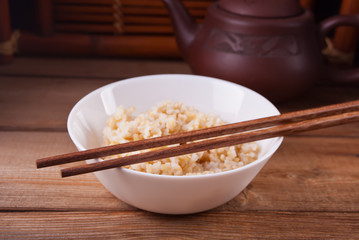 Cooked brown rice in white bowl with chopsticks on the wooden background.
