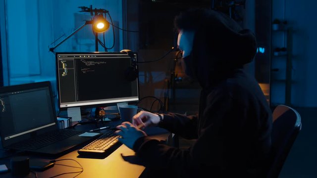 cybercrime, hacking and technology concept - asian male hacker in mask using computer for cyber attack and taking notes in dark room at night