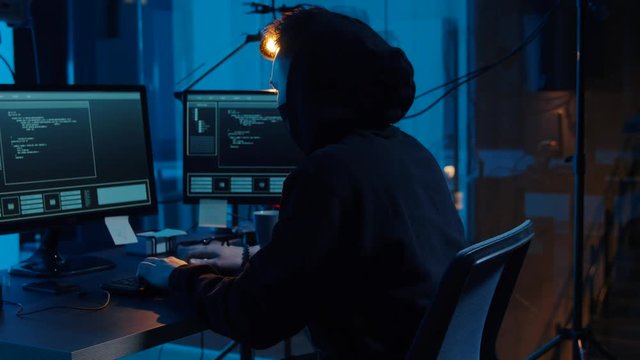 cybercrime, hacking and technology concept - asian male hacker in mask writing code or using computer virus program for cyber attack in dark room at night