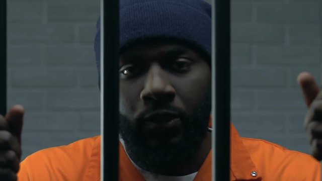 Black criminal aggressively holding prison bars and looking at camera, guilty