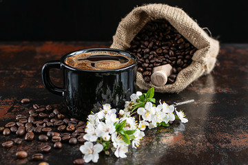 Composition artistique avec tasse de café aromatique chaud, brindille de cerise en fleurs et grains de café torréfiés sur fond sombre vintage.