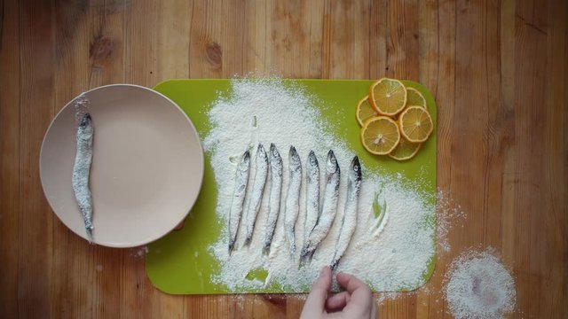Top view of a man's hands covering a capelin with flour on a wooden table, the process of making a small silver fish, preparation for frying in oil in a pan.