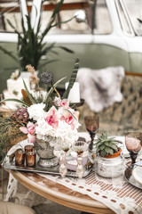Beautiful glasses, candles, flowers stand on a fruitful decorated table in rustic boho style. A large green sofa and hippy bus on the background. Summer desert atmosphere. Wedding party